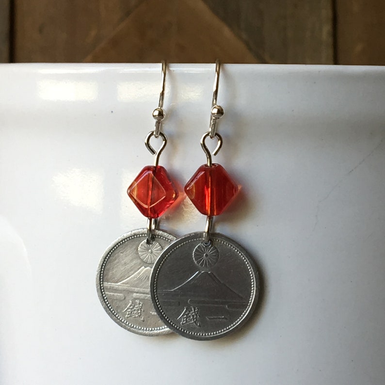May include: Silver earrings with red glass beads and Japanese coins featuring Mount Fuji. The coins have Japanese characters on them.