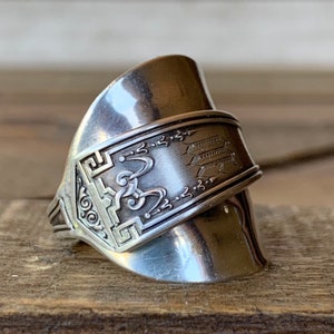 May include: A silver spoon ring with an ornate design and a monogram. The ring is made from a bent spoon and has a unique, handcrafted look.