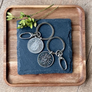 May include: Two vintage Dutch coin keychains with silver-colored metal rings and clasps. The coins are attached to a black slate square, which is placed on a wooden tray. Green sprigs are placed on the tray.