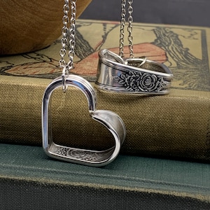 May include: Two silver heart-shaped pendants with floral designs hanging on chains. The pendants are made from repurposed silverware.