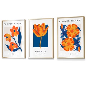 May include: Three framed prints with floral designs. The first print features a single orange and yellow tulip on a blue background with the text "BOTANICA PARIS - FRANCE". The second print features a branch of orange and yellow flowers with blue leaves on a cream background with the text "FLOWER MARKET". The third print features two orange and yellow flowers with blue leaves on a cream background with the text "FLOWER MARKET".