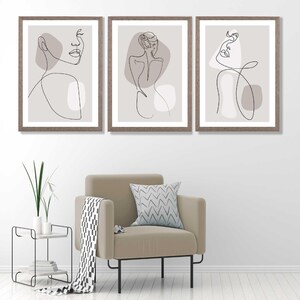 May include: Set of three framed prints featuring minimalist line drawings of female figures in various poses. The drawings are in black on a light grey background. The frames are a light brown wood.