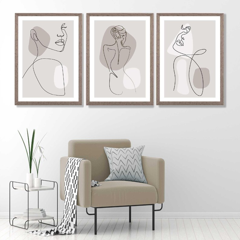 May include: Set of three framed prints featuring minimalist line drawings of female figures in various poses. The drawings are in black on a light grey background. The frames are a light brown wood.