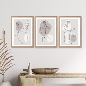 May include: Set of three minimalist line art prints featuring abstract figures with muted pink and grey tones. The prints are framed in natural wood.