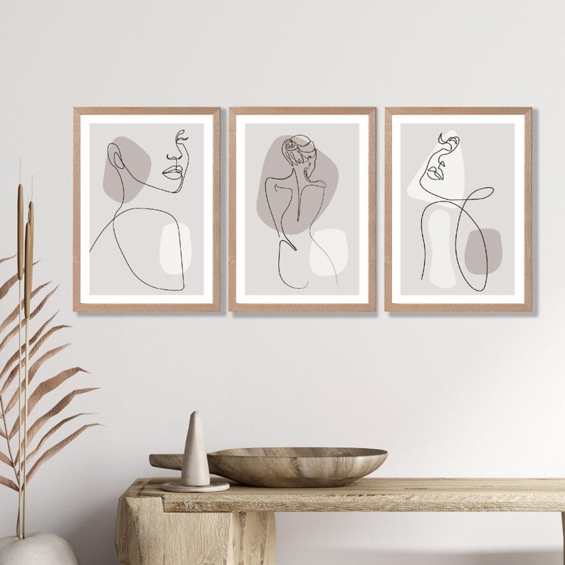May include: Set of three minimalist line art prints featuring abstract figures with muted pink and grey tones. The prints are framed in natural wood.
