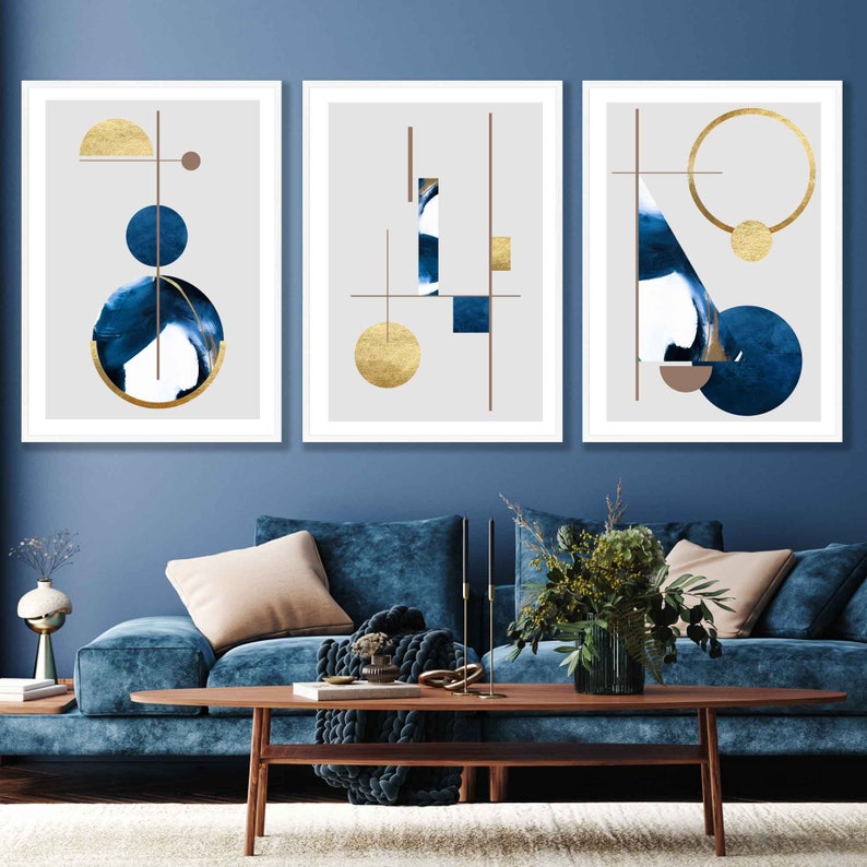 May include: Three framed abstract art prints with geometric shapes in shades of blue and gold on a gray background. The prints are hung on a blue wall.