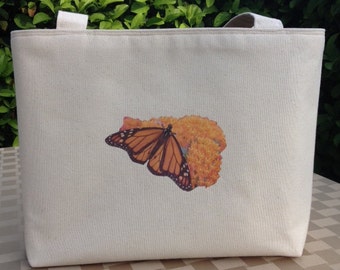 Puede incluir: Bolso tote de lona color crema con un diseño de mariposa monarca. La mariposa, con alas naranjas y negras, se posa sobre una flor naranja. El bolso tiene dos asas color crema y está hecho de lona resistente.