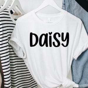 May include: A white t-shirt with the word "Daisy" in bold, black letters. The shirt is hanging on a white hanger, with other clothing items visible in the background. The shirt has short sleeves and a round neckline.