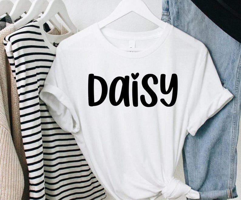 May include: A white t-shirt with the word "Daisy" in bold, black letters. The shirt is hanging on a white hanger, with other clothing items visible in the background. The shirt has short sleeves and a round neckline.
