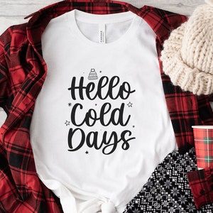 May include: White t-shirt with the text "Hello Cold Days" in black script, adorned with a winter hat and star graphics. The shirt is paired with a red and black plaid shirt, a cream-colored knit hat, and a plaid cup.