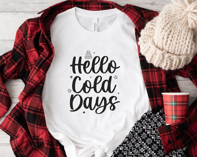 May include: White t-shirt with the text "Hello Cold Days" in black script, adorned with a winter hat and star graphics. The shirt is paired with a red and black plaid shirt, a cream-colored knit hat, and a plaid cup.