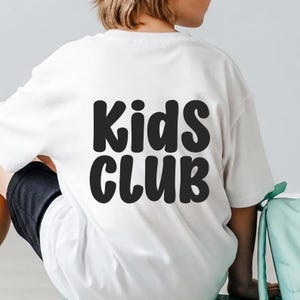 Puede incluir: Camiseta blanca con la inscripci&oacute;n "Kids CLUB" en letras negras grandes. La camiseta la lleva puesta una persona, con la espalda de la camiseta y parte del pantal&oacute;n corto visibles. La camiseta parece estar hecha de una tela suave.