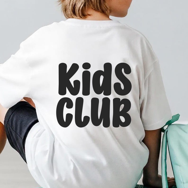 Puede incluir: Camiseta blanca con la inscripci&oacute;n "Kids CLUB" en letras negras grandes. La camiseta la lleva puesta una persona, con la espalda de la camiseta y parte del pantal&oacute;n corto visibles. La camiseta parece estar hecha de una tela suave.