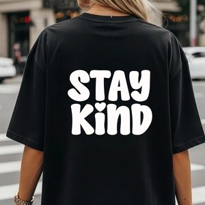 May include: Black t-shirt with the words "STAY KIND" in large, white, rounded letters. A small heart graphic is between the words. The shirt is made of a soft material.