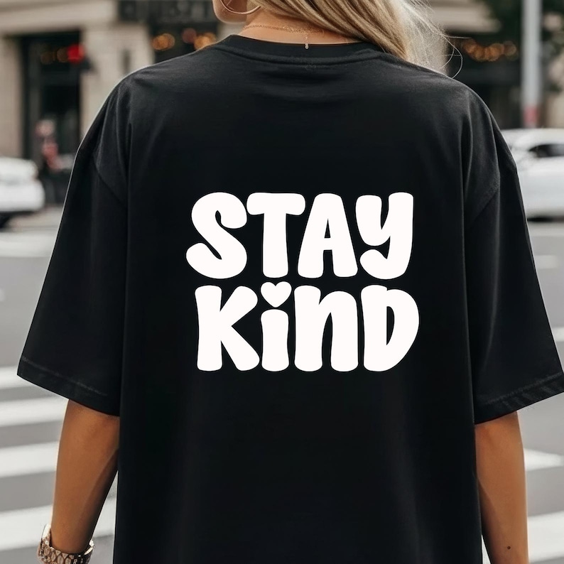 May include: Black t-shirt with the words "STAY KIND" in large, white, rounded letters. A small heart graphic is between the words. The shirt is made of a soft material.