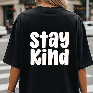 Puede incluir: Camiseta negra con las palabras "Stay Kind" en letras grandes, blancas y redondeadas. La camiseta est&aacute; hecha de un material suave y tiene mangas cortas. La imagen est&aacute; tomada desde atr&aacute;s, mostrando el texto de forma prominente.