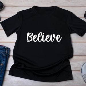 May include: A black t-shirt with the word "Believe" in white script across the front. The shirt is short-sleeved and appears to be made of a soft material. The shirt is displayed on a light-colored wooden surface.