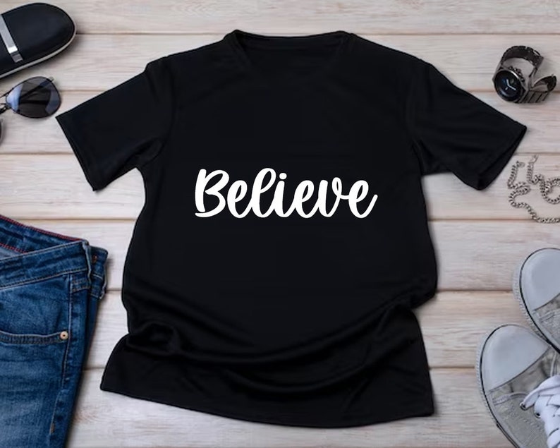 May include: A black t-shirt with the word "Believe" in white script across the front. The shirt is short-sleeved and appears to be made of a soft material. The shirt is displayed on a light-colored wooden surface.