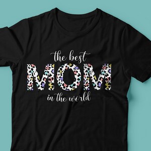 Puede incluir: Camiseta negra con el texto "the best MOM in the world" en letras blancas. La palabra "MOM" est&aacute; en un estampado de leopardo colorido.