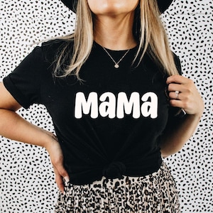 Puede incluir: Camiseta negra con la palabra blanca "Mama" impresa en la parte delantera. La camiseta tiene mangas cortas y est&aacute; anudada a la cintura. El fondo es un estampado de lunares en blanco y negro.