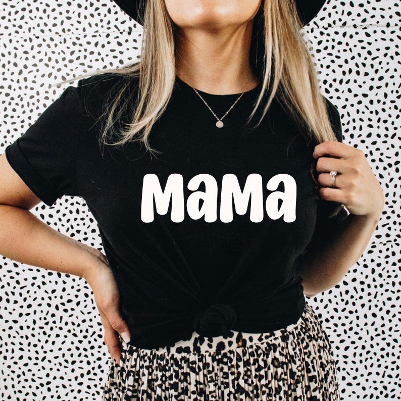 Puede incluir: Camiseta negra con la palabra blanca "Mama" impresa en la parte delantera. La camiseta tiene mangas cortas y est&aacute; anudada a la cintura. El fondo es un estampado de lunares en blanco y negro.