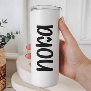 May include: A white tumbler with a clear lid, featuring the name "Nora" in bold black letters. The tumbler is held by a person's hand, showcasing the personalized design. The background includes a decorative plant and a white surface.