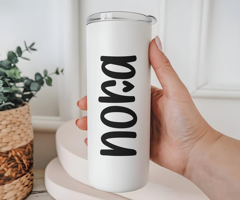 May include: A white tumbler with a clear lid, featuring the name "Nora" in bold black letters. The tumbler is held by a person's hand, showcasing the personalized design. The background includes a decorative plant and a white surface.