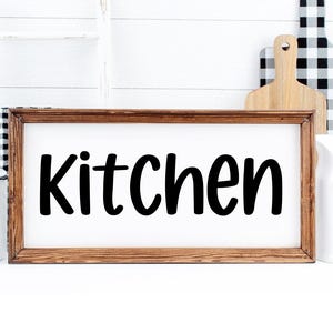 May include: A framed wooden sign with the word "Kitchen" in bold, black lettering. The sign is set against a white background, with a white pitcher, a wooden cutting board, and a black and white checkered cutting board in the background.