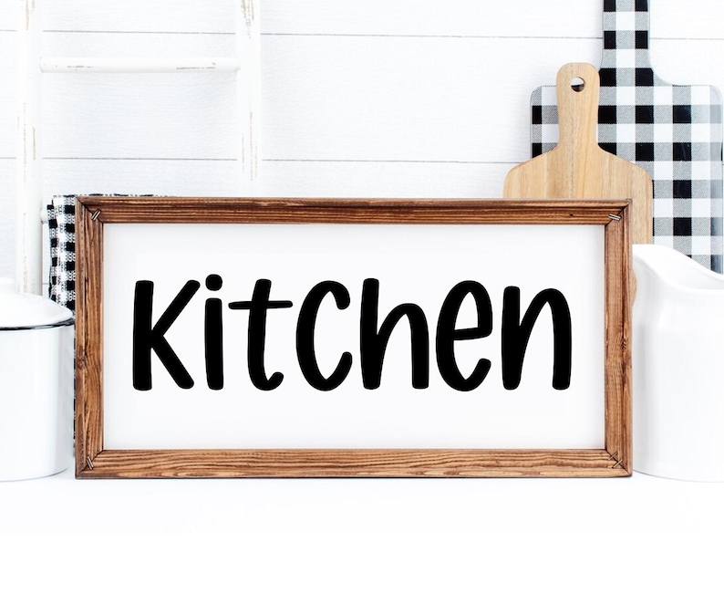 May include: A framed wooden sign with the word "Kitchen" in bold, black lettering. The sign is set against a white background, with a white pitcher, a wooden cutting board, and a black and white checkered cutting board in the background.