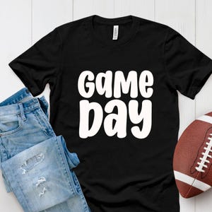 Puede incluir: Camiseta negra con las palabras "GAME DAY" en blanco, en negrita y redondeadas. Un par de vaqueros azules y un bal&oacute;n de f&uacute;tbol marr&oacute;n tambi&eacute;n est&aacute;n en la imagen. La camiseta es de un material suave.