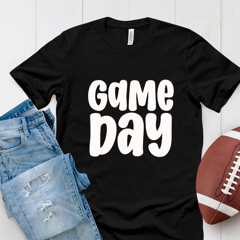 Puede incluir: Camiseta negra con las palabras "GAME DAY" en blanco, en negrita y redondeadas. Un par de vaqueros azules y un bal&oacute;n de f&uacute;tbol marr&oacute;n tambi&eacute;n est&aacute;n en la imagen. La camiseta es de un material suave.