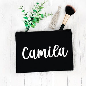 May include: Black cosmetic bag with the name "Camila" in white script. The bag is open, revealing a makeup brush, a hair tie, and a sprig of greenery. The bag is on a white wooden surface.