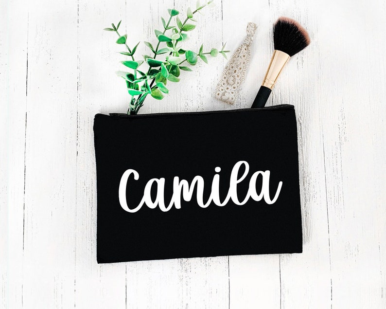 May include: Black cosmetic bag with the name "Camila" in white script. The bag is open, revealing a makeup brush, a hair tie, and a sprig of greenery. The bag is on a white wooden surface.