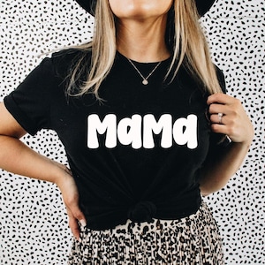 May include: Black t-shirt with the word "Mama" in white, bold, rounded letters. The shirt is knotted at the waist. The background is a black and white spotted pattern. The model is wearing a necklace and a ring.
