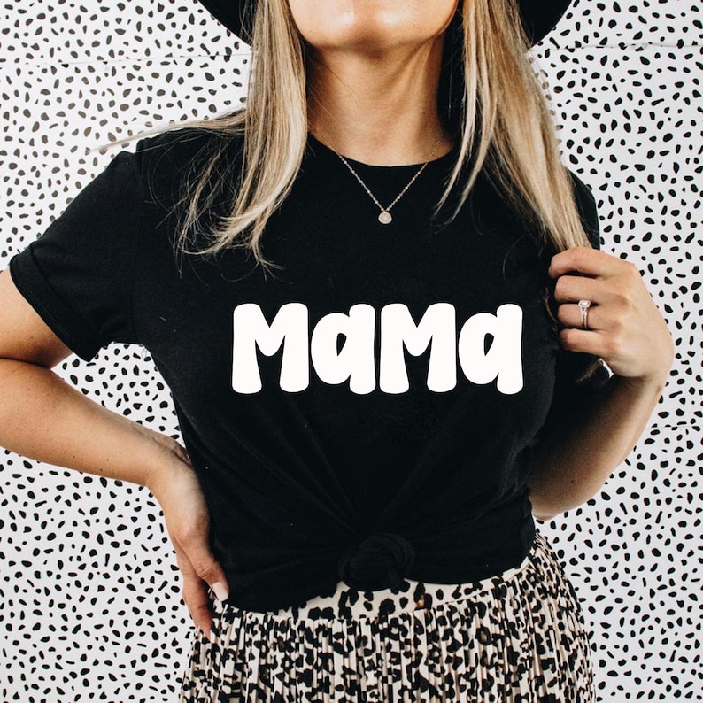 May include: Black t-shirt with the word "Mama" in white, bold, rounded letters. The shirt is knotted at the waist. The background is a black and white spotted pattern. The model is wearing a necklace and a ring.