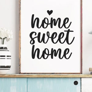 May include: A framed print with the words "home sweet home" in black script, topped with a heart. A vase with white flowers and a black and white striped design sits to the left. The print is on a shelf with a blue painted surface.