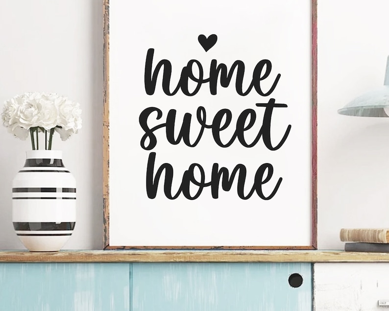 May include: A framed print with the words "home sweet home" in black script, topped with a heart. A vase with white flowers and a black and white striped design sits to the left. The print is on a shelf with a blue painted surface.