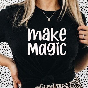 May include: Black t-shirt with the words "make magic" in white script. The shirt is knotted at the waist. The background is a black and white spotted pattern.