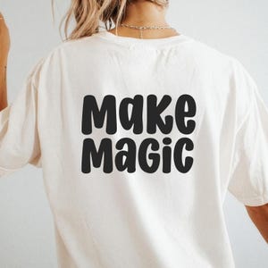 Puede incluir: Camiseta color crema con las palabras "Make Magic" impresas en letras negras y en negrita. La camiseta est&aacute; hecha de un material suave y tiene mangas cortas. El texto est&aacute; centrado en la parte posterior de la camiseta.