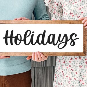 May include: A rectangular wooden sign with the word "Holidays" in black cursive lettering. The sign is held by two people, with a light brown wooden frame and a white background. The sign is a decorative item.