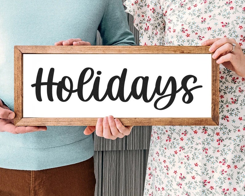 May include: A rectangular wooden sign with the word "Holidays" in black cursive lettering. The sign is held by two people, with a light brown wooden frame and a white background. The sign is a decorative item.