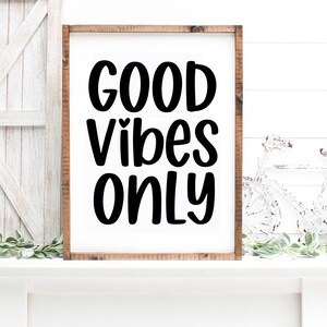 May include: A framed wooden sign with the words "GOOD VIBES ONLY" in bold, black lettering. The sign is set against a white background and rests on a white surface. Decorative greenery and a white bicycle are visible.