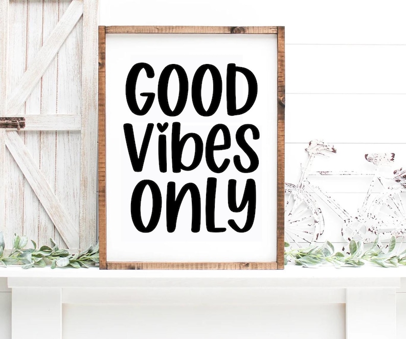 May include: A framed wooden sign with the words "GOOD VIBES ONLY" in bold, black lettering. The sign is set against a white background and rests on a white surface. Decorative greenery and a white bicycle are visible.