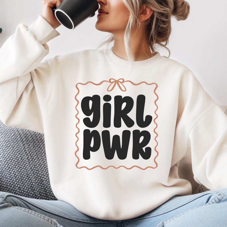 Puede incluir: Sudadera color crema con las palabras "GIRL PWR" en negro, dentro de un marco decorativo marr&oacute;n. La sudadera tiene mangas largas y cuello redondo. El texto est&aacute; en negrita y con una fuente redondeada.