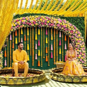 May include: A vibrant outdoor wedding ceremony setup with yellow drapery, floral arrangements, and decorative accents. Two people are seated in ornate, golden-colored bowls. The backdrop features a floral arch and colorful hanging tassels.