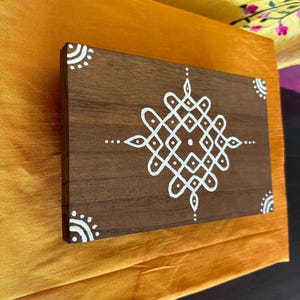Op de afbeelding: Een bruine houten plaquette met een wit geometrisch ontwerp erop geschilderd. Het ontwerp is een traditioneel Indiaas rangoli-patroon.