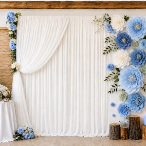 May include: A white fabric backdrop featuring a vertical arrangement of blue and white paper flowers on the right side and a smaller floral cluster on the left. The display includes a white draped curtain, wooden logs, and a tall metal lantern.