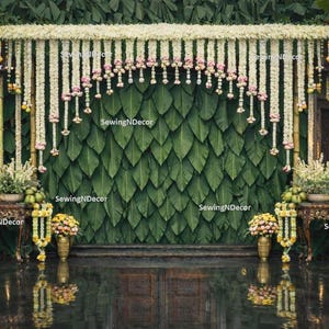 May include: A decorative backdrop featuring a wall of large green leaves, adorned with floral garlands and hanging decorations. The scene includes ornate tables with floral arrangements and a reflective floor, creating an elegant setting.