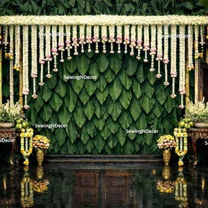 May include: A decorative event backdrop featuring a wall of layered green leaves, adorned with hanging white and pink floral garlands. Two wooden tables flank the center, each holding floral arrangements and yellow fruit, with the text SewingNDecor.