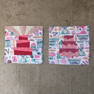 May include: Two square fabric pieces with a patchwork design. One features a fan-like pattern and pink and red blocks. The other has a repeating pattern of kitchen tools and pink blocks. Both are on a concrete surface.